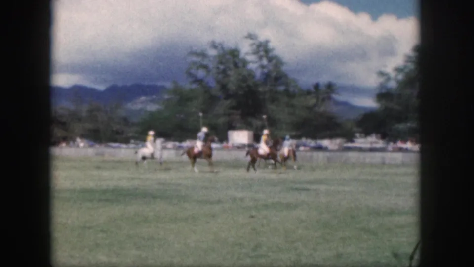 Distant mounted polo players in white and yellow uniforms ride across an Oahu field, with trees and mountains beyond.