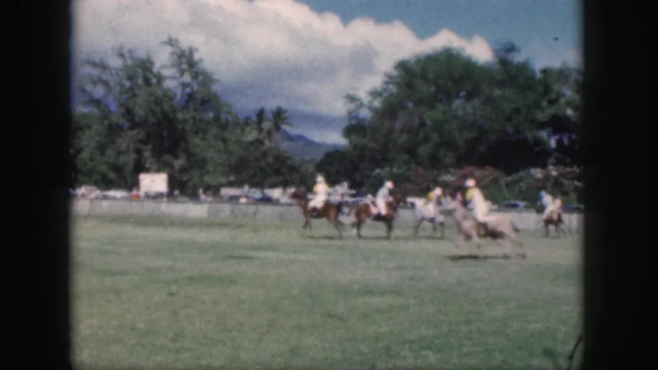 Final Oahu polo frame showing riders moving to the right side of the grassy field, with a pale horse, trees, and clouds visible.