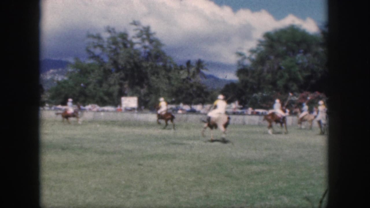 1959 Oahu home-movie frame showing polo players on horseback moving across a field with trees and mountains in the distance.