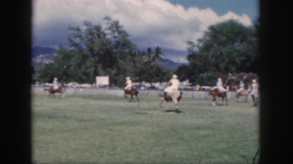 1959 Oahu home-movie frame showing polo players on horseback moving across a field with trees and mountains in the distance.