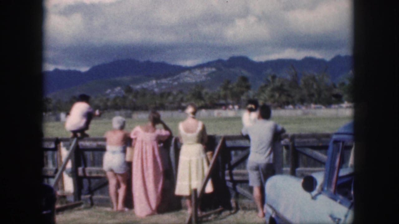 Spectators in 1950s clothing watch a polo match from behind a wooden fence in Oahu, with mountains, palm trees, and a blue car nearby.