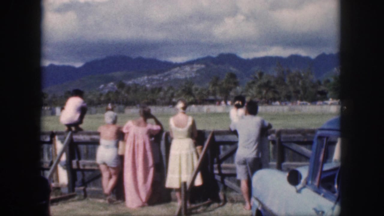 1959 Oahu home-movie frame with women and other spectators at a paddock fence, mountains and palms beyond.