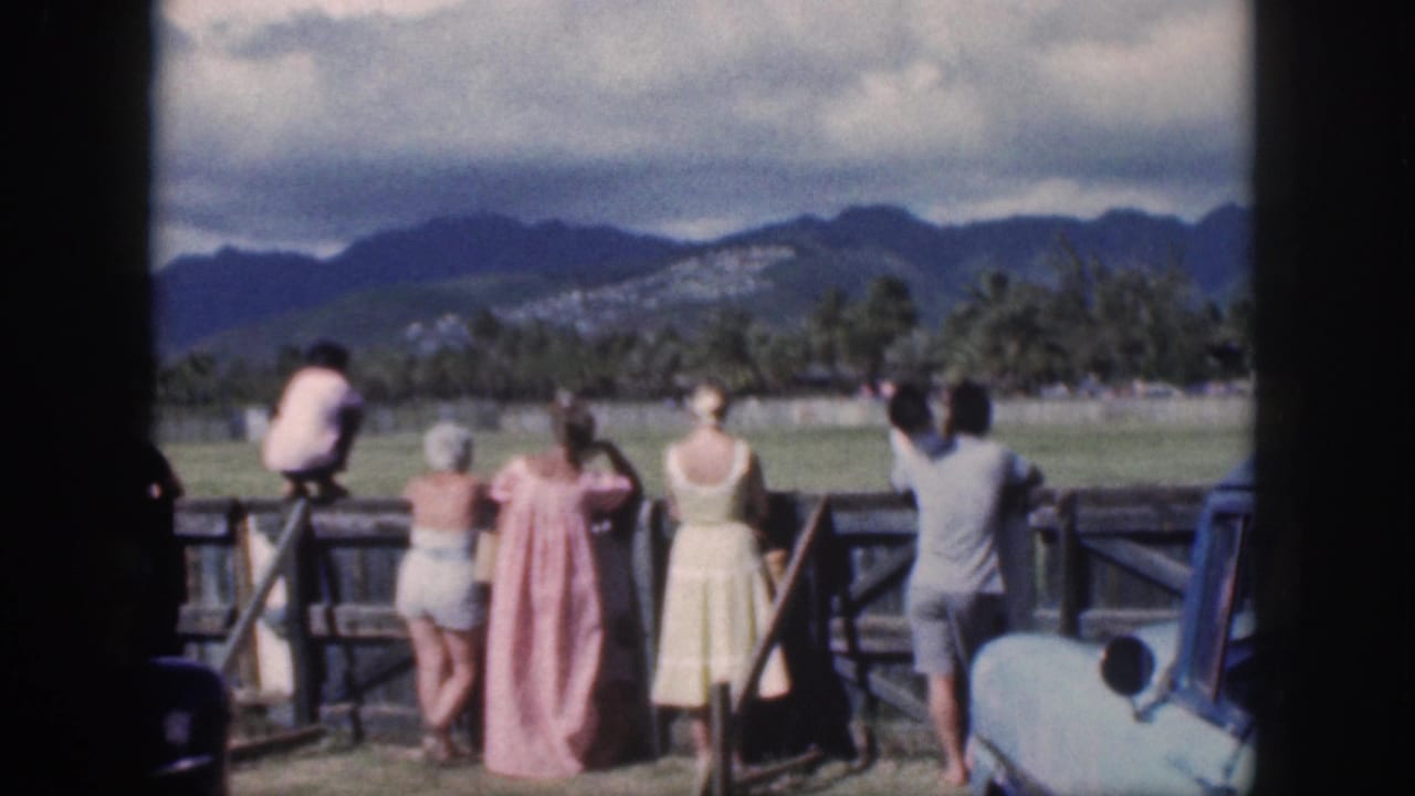 Spectators in mid-century clothing watch horseback activity in an Oahu paddock, with palms and mountains in the background.