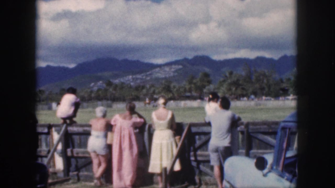 Oahu paddock scene showing spectators from behind, a distant rider, palms, mountains, and a partial blue car at right.