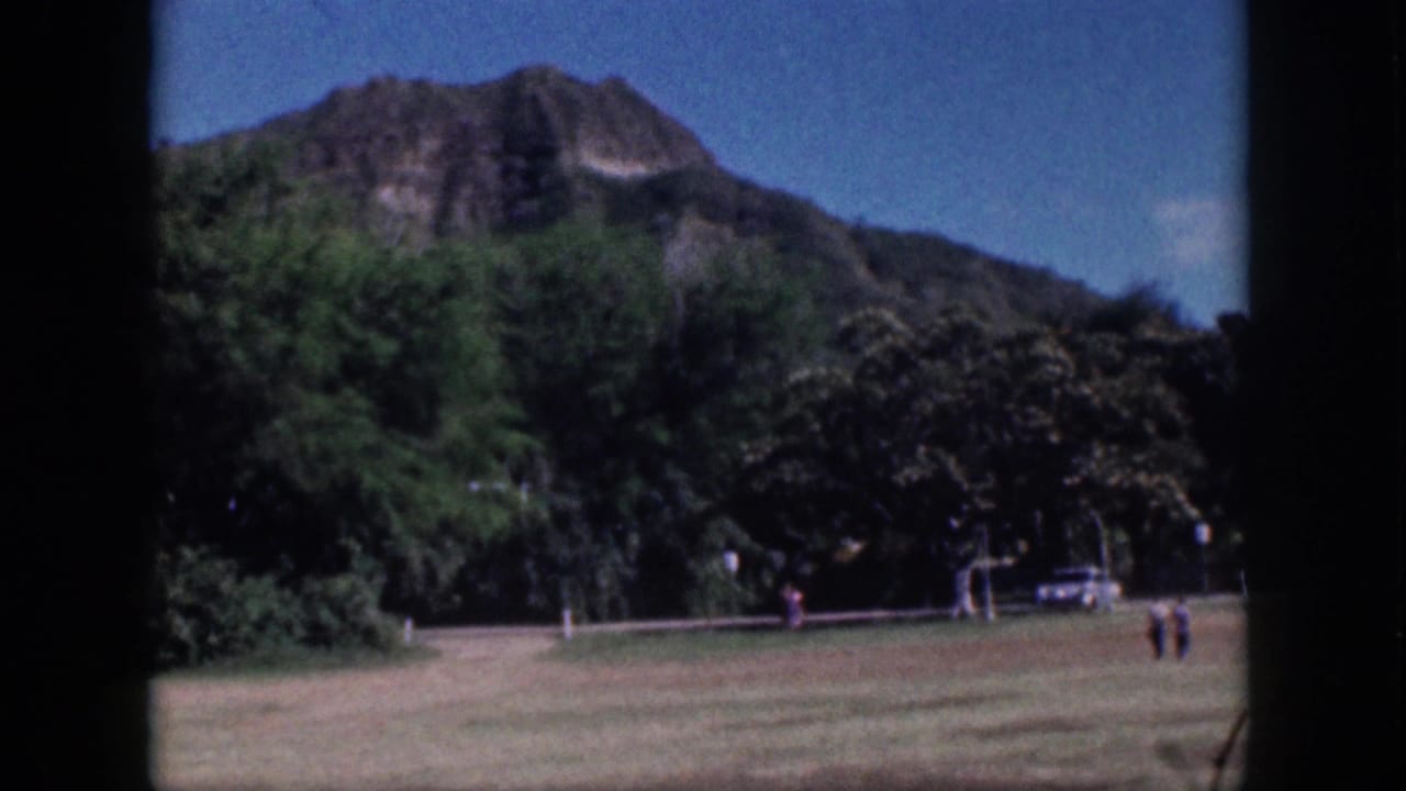 1959 Oahu travel frame showing a broad grassy foreground, dense trees, and a mountain slope under blue sky.