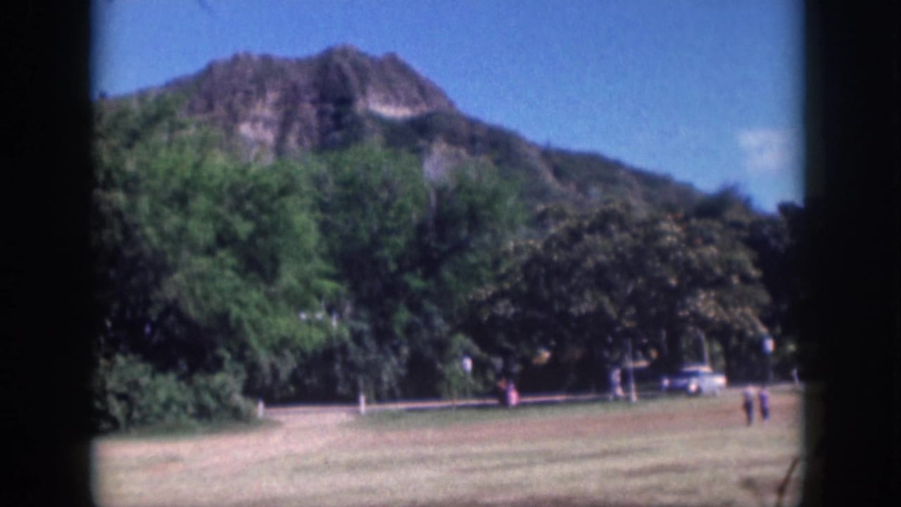 1959 Oahu home-movie frame showing a mountain ridge beyond trees, with a grassy field, roadside cars, and distant walkers.