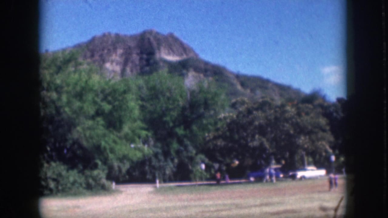 1959 Oahu frame with a mountain backdrop, dense trees, open grass, a roadside white car, and small figures walking nearby.