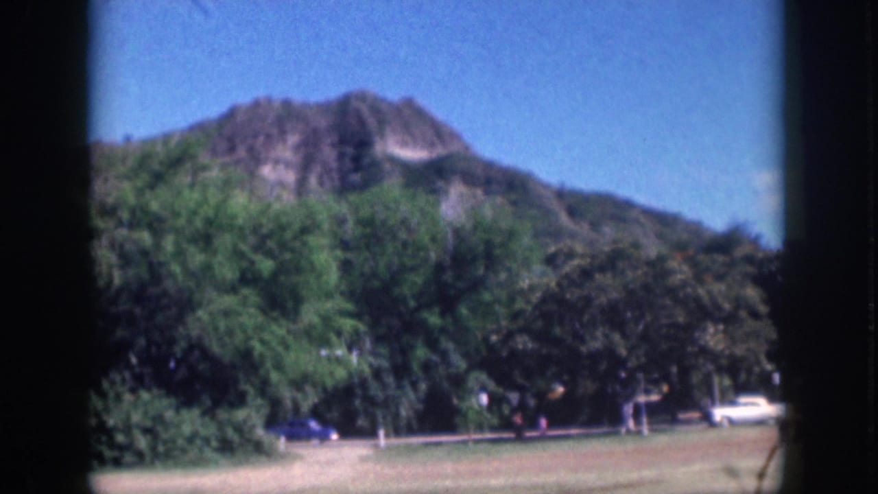 1959 Oahu home-movie frame showing trees, shrubs, grass, a mountain ridge, roadside cars, and distant pedestrians.