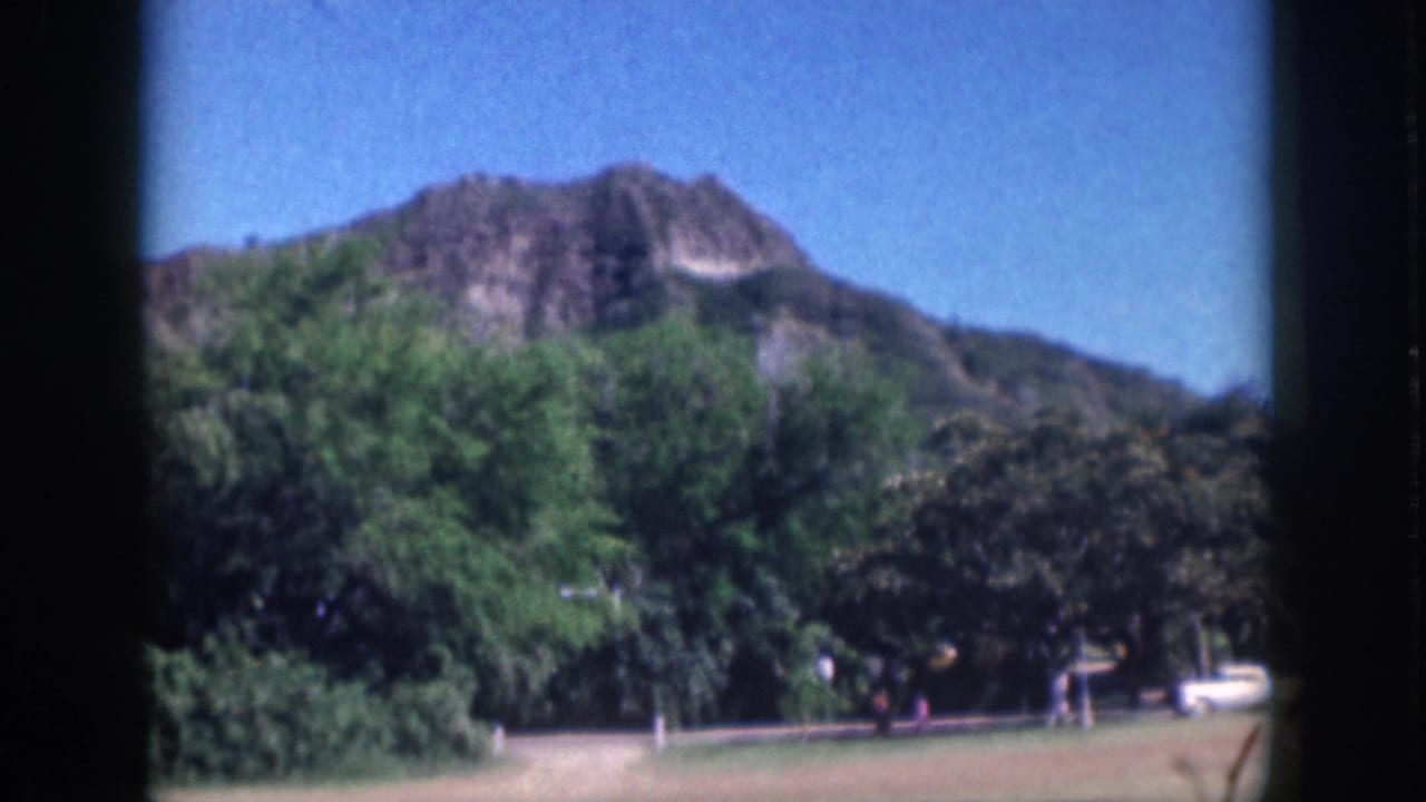 Soft 1959 Oahu 8mm frame with a mountain ridge above trees, open grass, a roadside white car, and distant people.