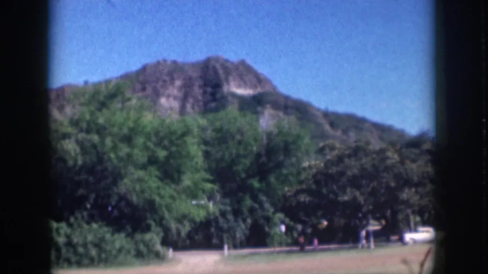 Soft 1959 Oahu 8mm frame with a mountain ridge above trees, open grass, a roadside white car, and distant people.