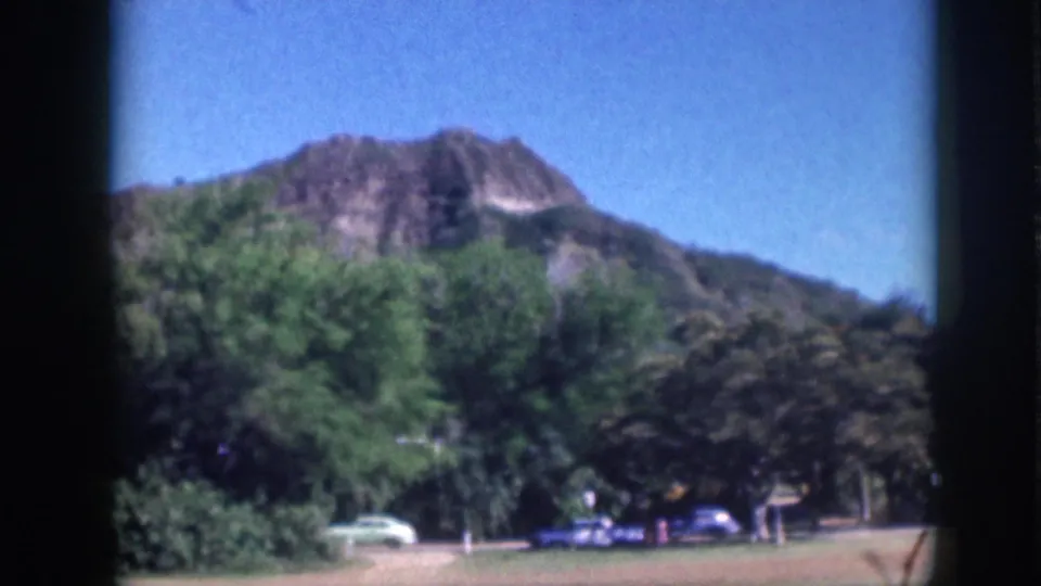 1959 Oahu footage showing a volcanic hillside beyond trees, with a white sedan and blue car near a park path.