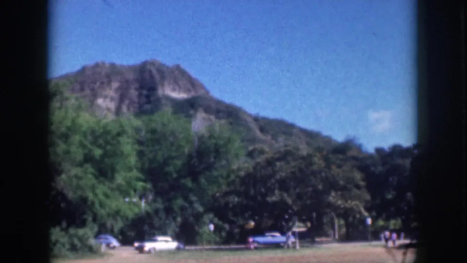 Oahu 1959 home-movie frame with distant people by a park path, cars, trees, and a Diamond Head-like hillside beyond.