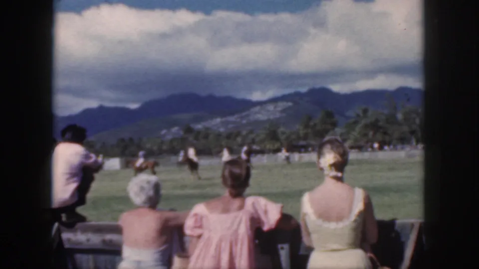 Oahu 1959 resort spectators view a polo match from behind a wooden rail with mountains and palms beyond the field.