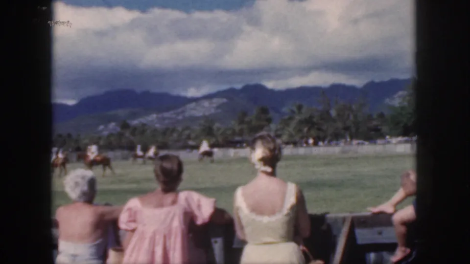 Oahu 1959 home-movie frame of women watching polo from a rail, with riders, palms, and dark mountain ridges beyond.