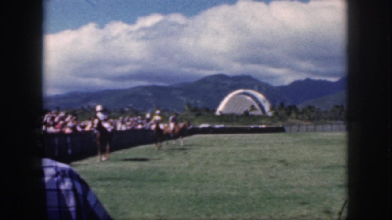 Polo riders on a grass field with spectators, mountains, clouds, and a white arch in the distance.