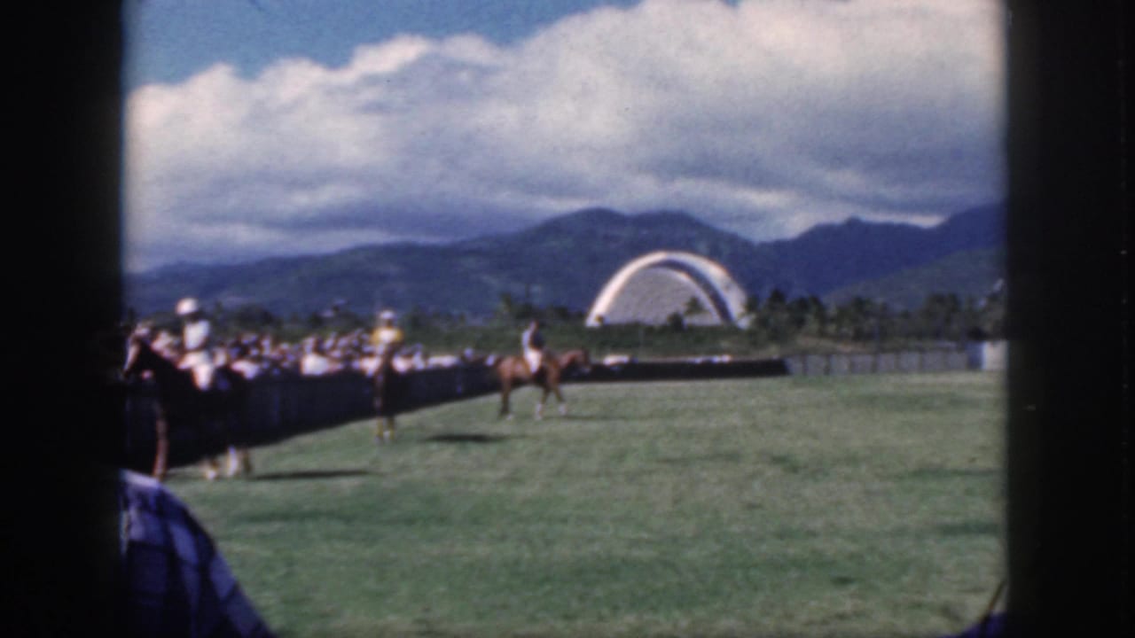 Two riders move across a polo field in front of a crowd, with mountains and a white arch behind them.