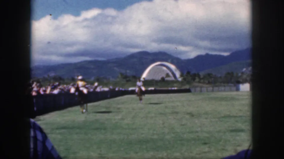 1959 Oahu home-movie view of a polo rider approaching the sideline crowd, with a white arched structure and mountains in the background.