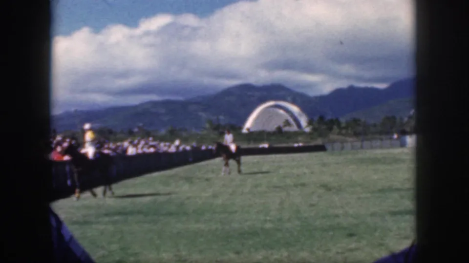 Oahu, 1959 frame of a polo horse and rider near spectators, with a green field, distant palms, mountains, clouds, and a white arch beyond.
