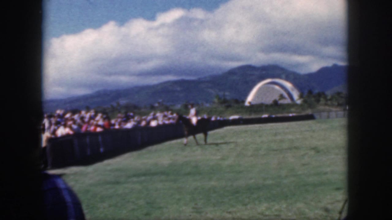 1959 Oahu footage frame with a mounted polo player near spectators, a field barrier, distant mountains, clouds, and a white arched structure.