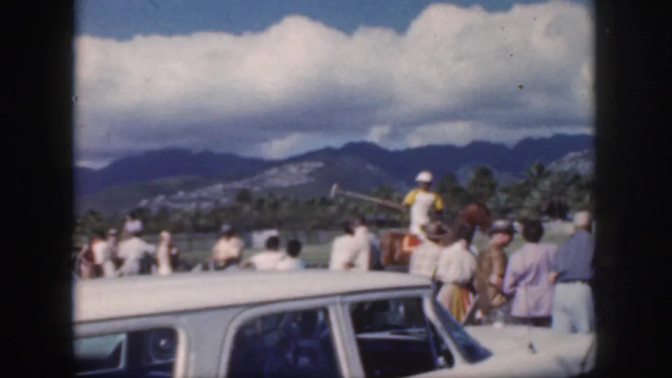 1959 Oahu home-movie view from behind spectators and a white car toward a polo rider, field, palm trees, mountains, and heavy clouds.