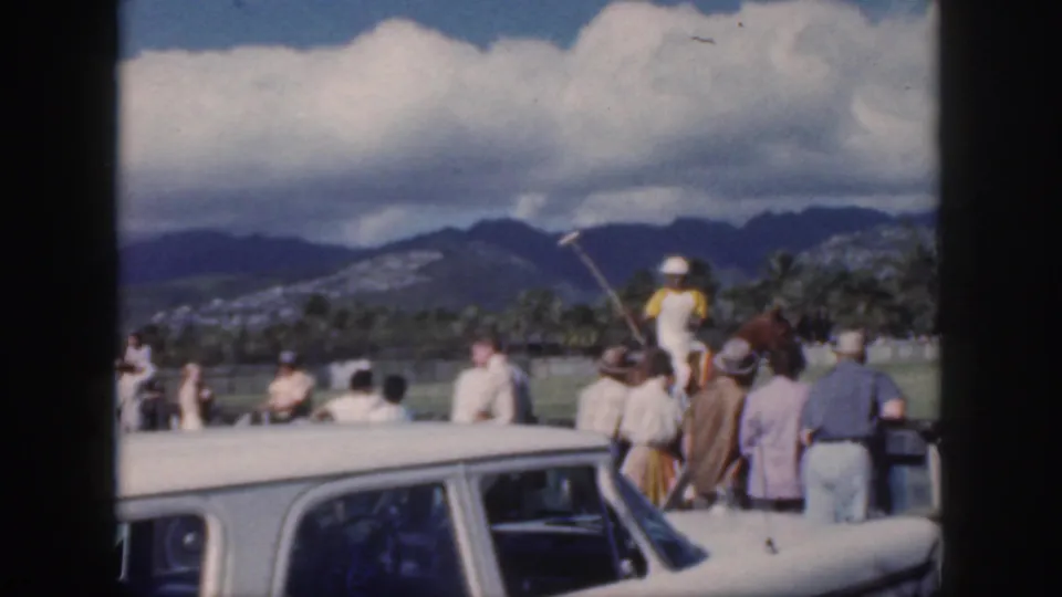 1959 Oahu home-movie frame showing spectators beside a white car watching a polo rider, with palms, mountains, and clouds beyond the field.