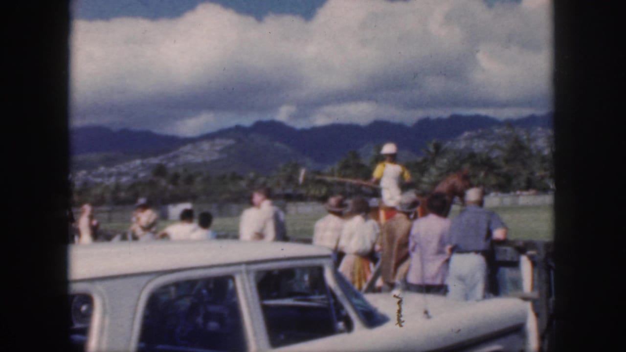 1959 Oahu home-movie frame showing a mounted polo player with mallet near spectators, a white sedan, palm trees, and mountains beyond the field.