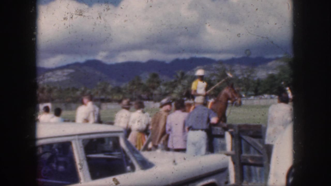 1959 Oahu frame of a polo player in a white helmet riding a brown horse near field spectators, with palm trees and mountains in the distance.