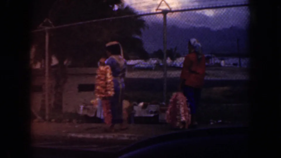 1959 Oahu frame with two women near lei displays by a chain-link fence, with cloudy mountains beyond and the scene partly bordered by a car window.
