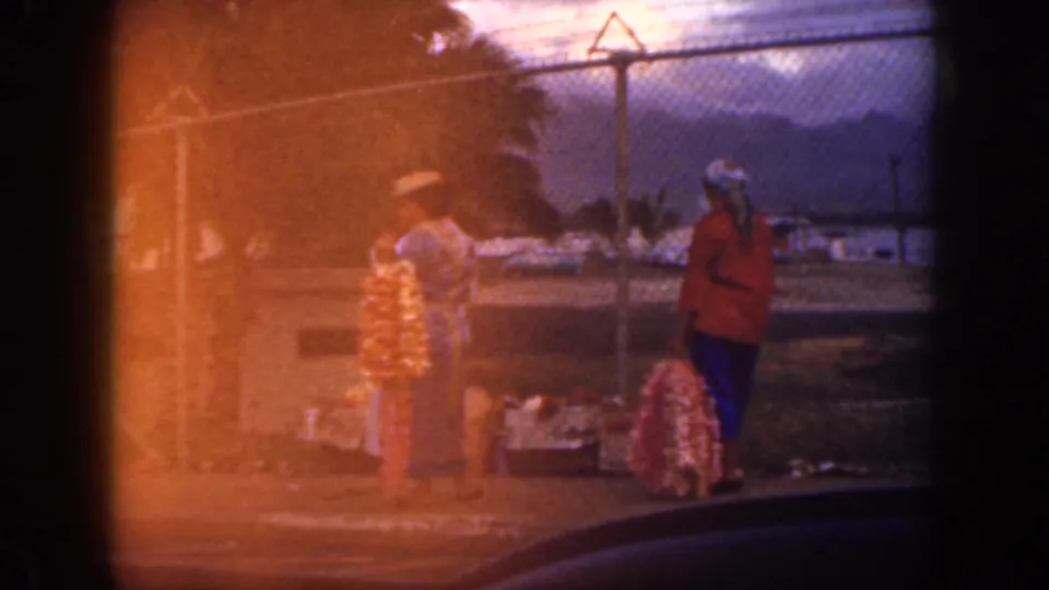 1959 Oahu home-movie frame where orange film flare partly covers two women, lei displays, a fence, palm foliage, and distant mountains.