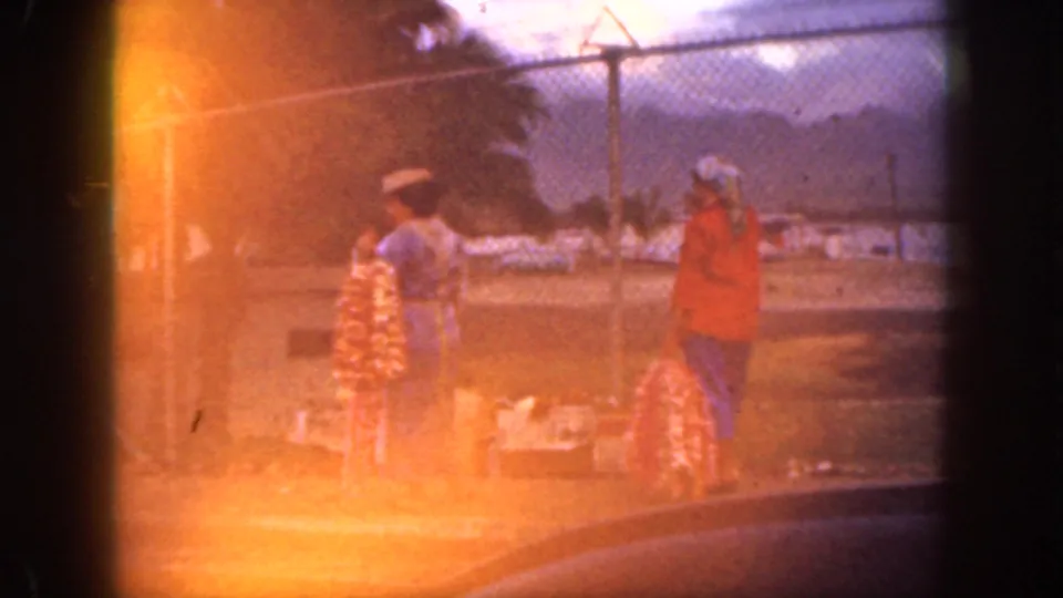 1959 Oahu frame with strong orange lens flare over two women by lei displays, a chain-link fence, palms, mountains, and a cloudy sky.
