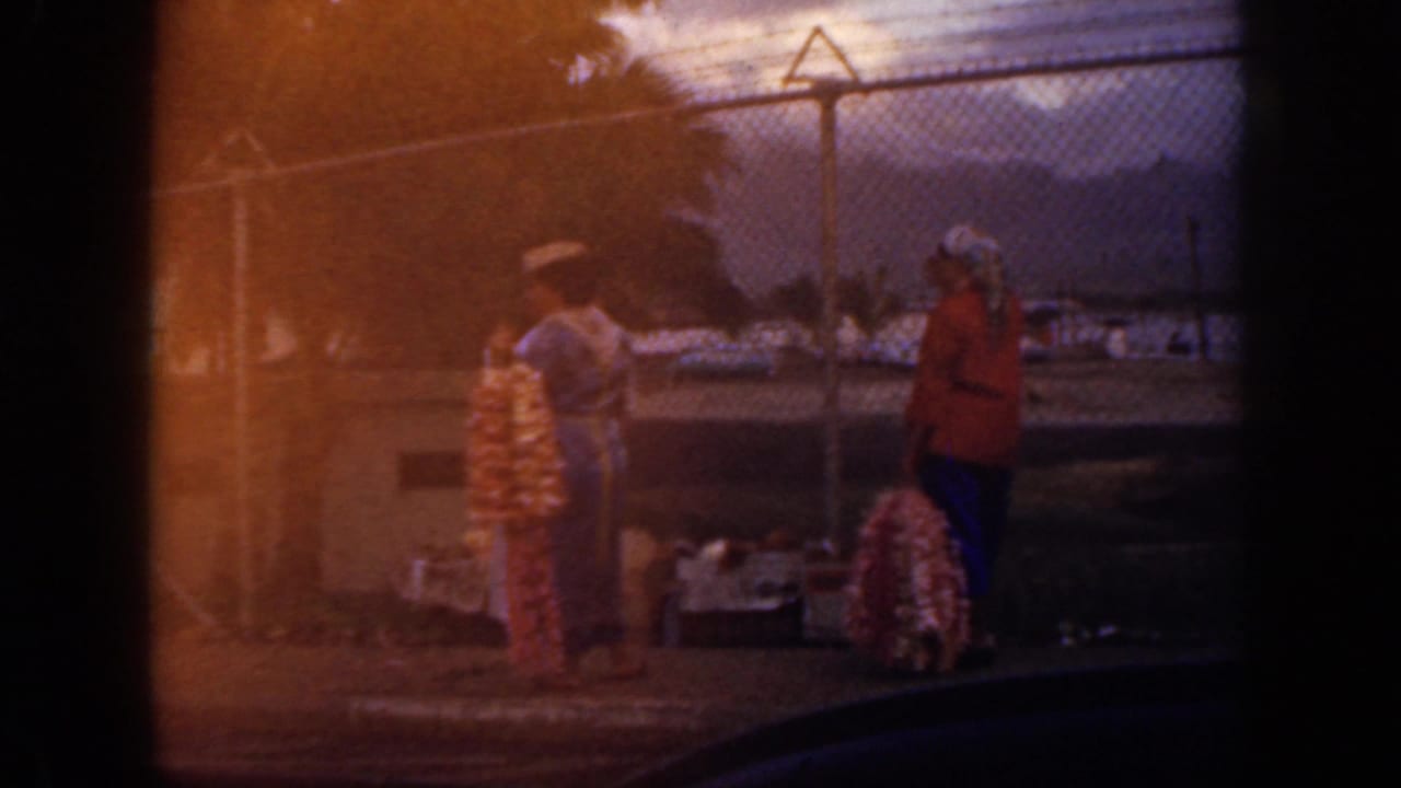 1959 Oahu home-movie frame showing two women near colorful leis beside a chain-link fence, with waterfront and mountains beyond.