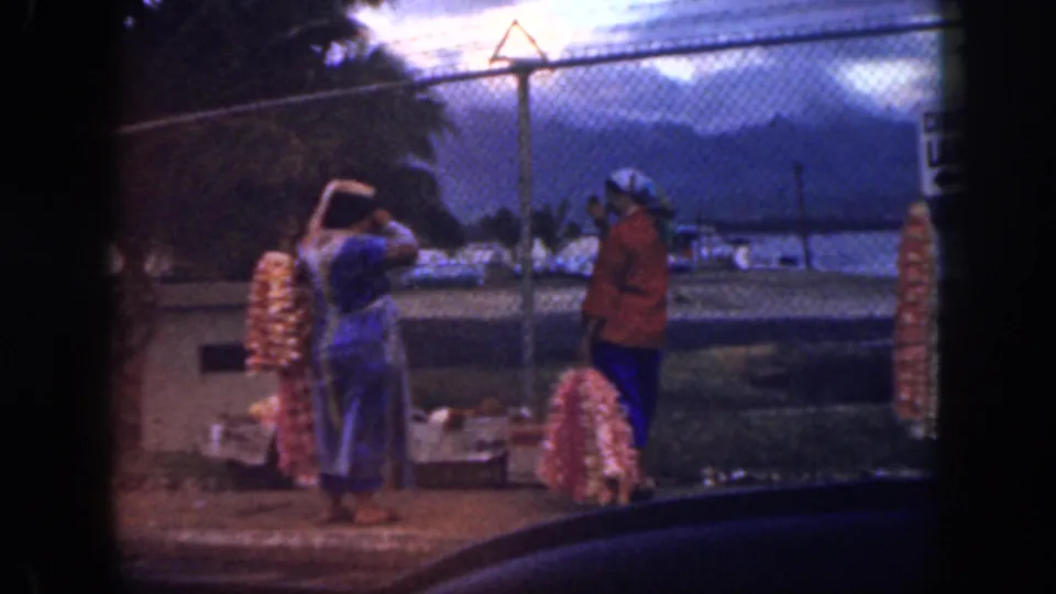 1959 Oahu home-movie frame of a woman in blue gesturing near another woman in red beside flower leis and a waterfront fence.