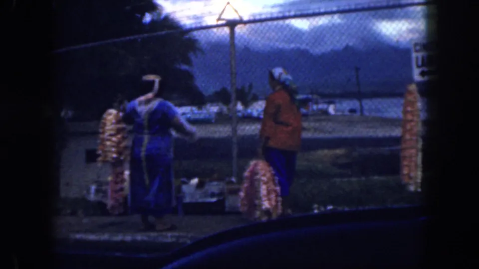 1959 Oahu frame showing two women facing each other near colorful leis, seen through dark car-window edges with water and mountains beyond.
