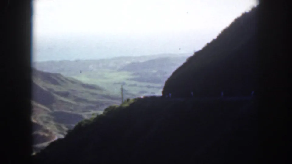 1959 Oahu travel frame looking from a shadowed tunnel area toward rolling green hills and a bright valley beyond.