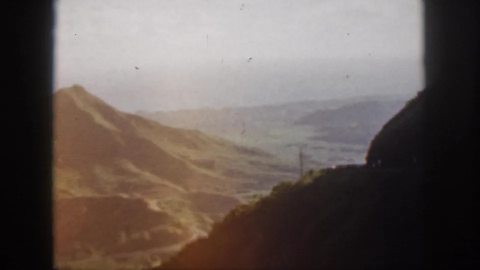 1959 Oahu frame showing sunlit mountain slopes, muted green valley land, and dark foreground hills from a moving road view.