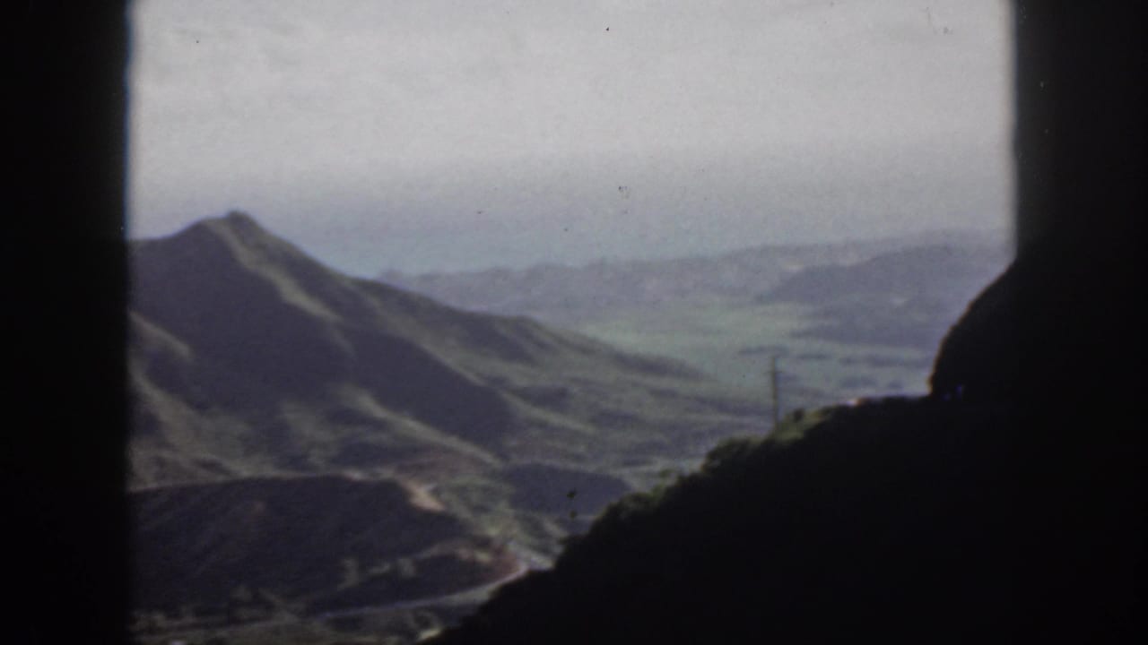 1959 Oahu home-movie frame showing grassy hills, a distant valley, and pale sky bordered by a dark window opening.
