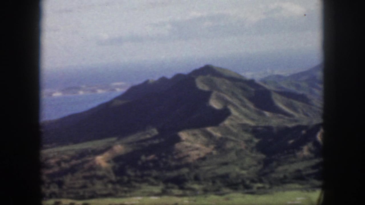 1959 Oahu home-movie frame showing a dark volcanic ridge, green slopes, blue ocean, and hazy cloud cover from an aerial viewpoint.