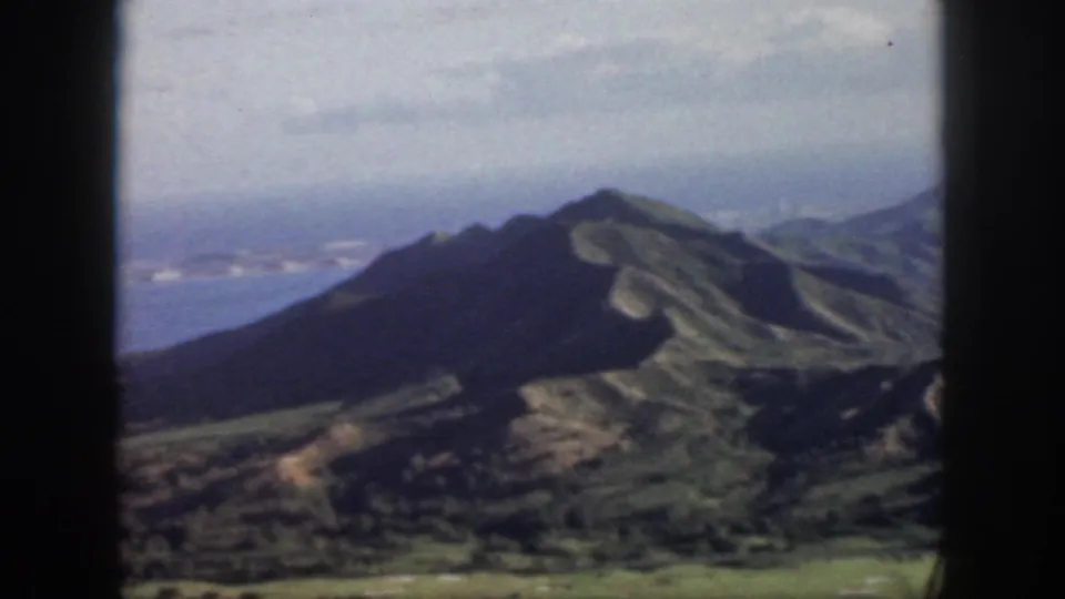 1959 Oahu home-movie frame showing a dark volcanic ridge, green slopes, blue ocean, and hazy cloud cover from an aerial viewpoint.