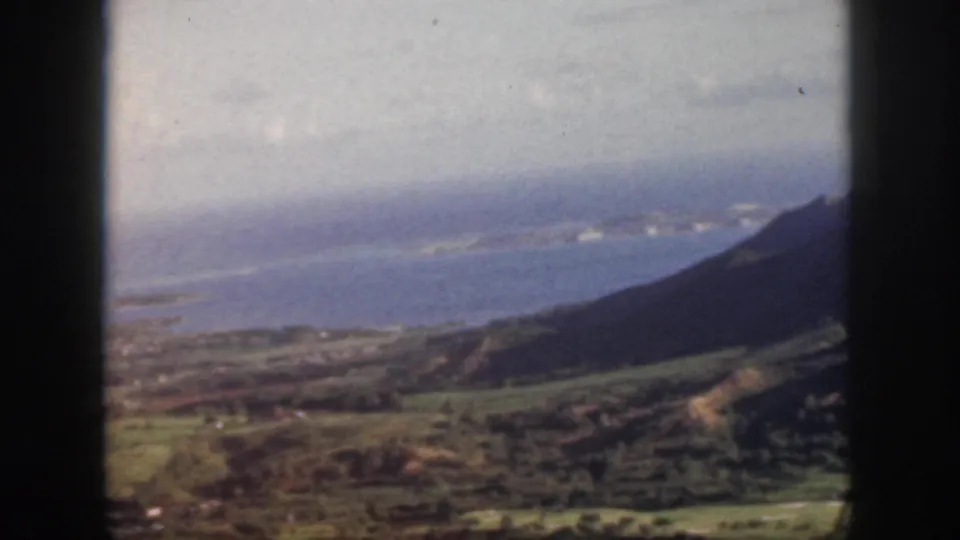 1959 Oahu 8mm frame showing a wider aerial look across green hills, blue coastal water, hazy clouds, and a dark ridge at the edge.