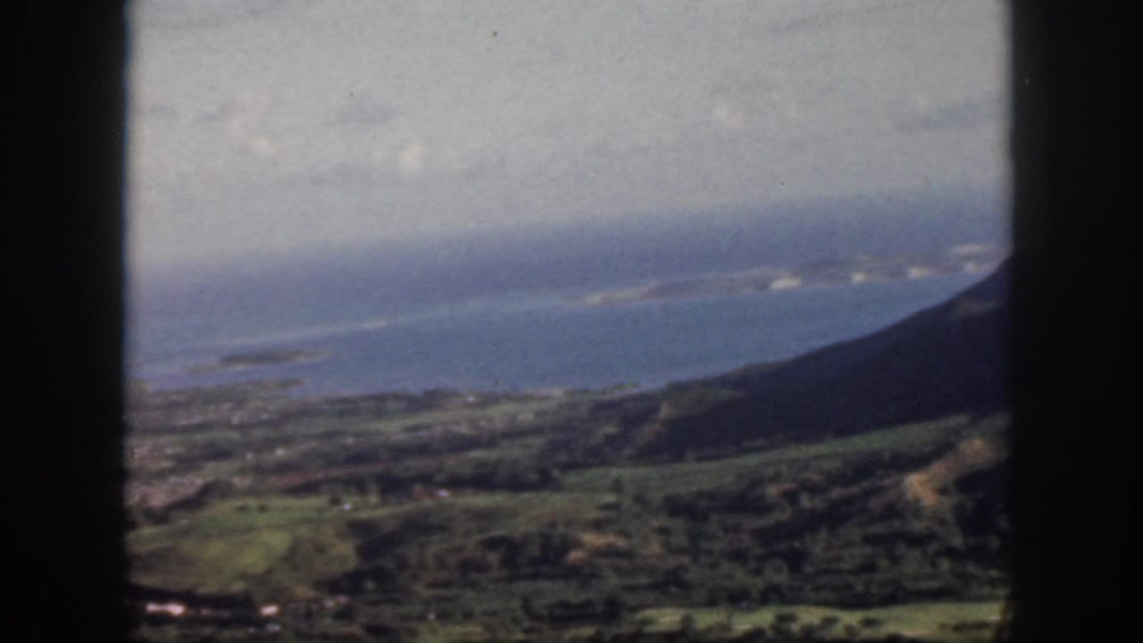 1959 Oahu home-movie aerial view showing green hills, coastal settlement, and blue Pacific water through a dark window frame.
