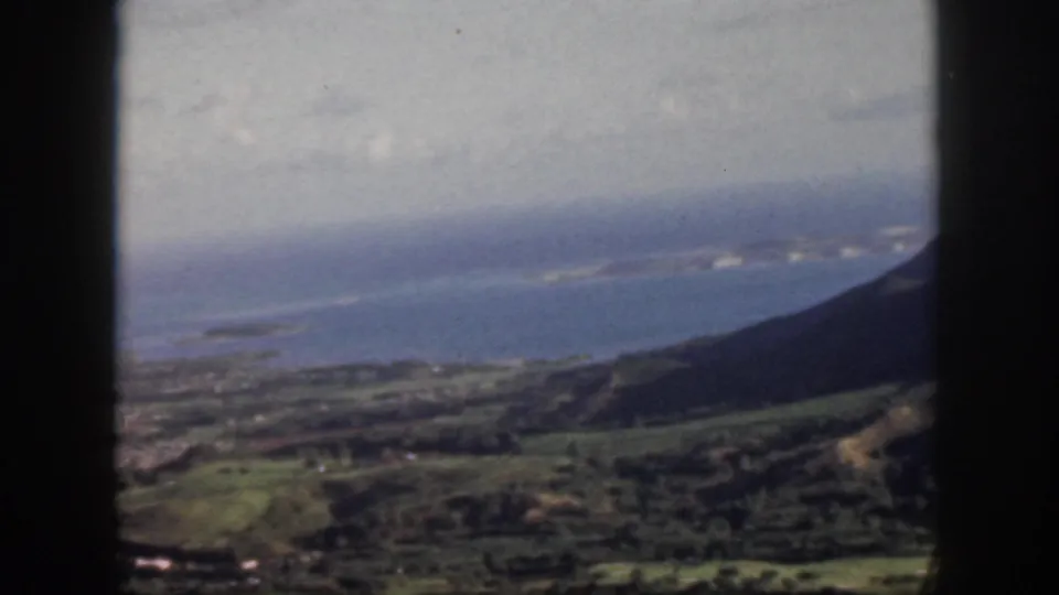 1959 Oahu home-movie aerial view showing green hills, coastal settlement, and blue Pacific water through a dark window frame.