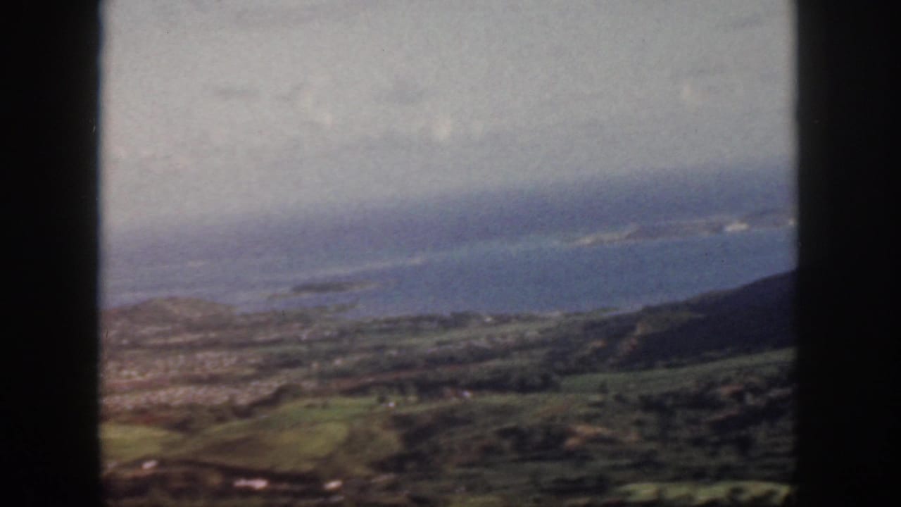 1959 Oahu aerial frame with blue Pacific water, coastal land, green slopes, and dark aircraft-window borders.