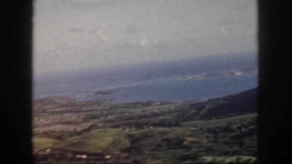 1959 Oahu aerial frame with blue Pacific water, coastal land, green slopes, and dark aircraft-window borders.