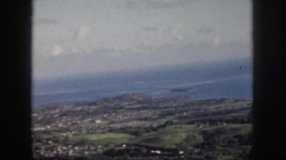 1959 Oahu aerial view with coastal settlement, green land, blue Pacific water, and distant offshore forms in haze.