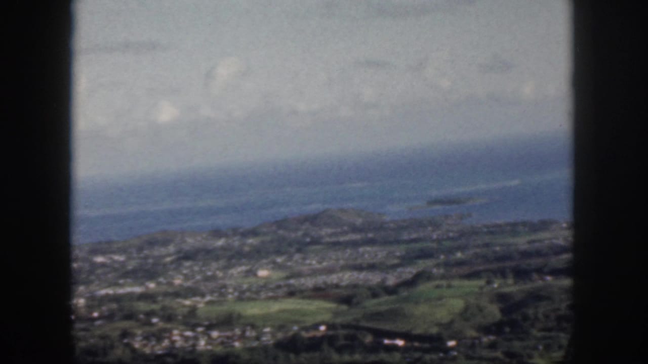 1959 Oahu home-movie frame showing a coastal town, green hills, ocean, and clouds through a dark window frame.