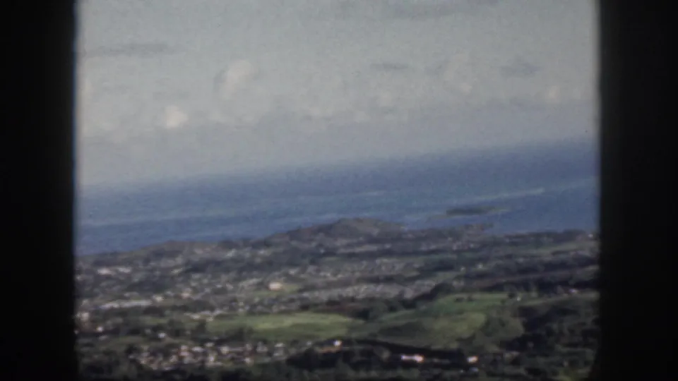1959 Oahu home-movie frame showing a coastal town, green hills, ocean, and clouds through a dark window frame.