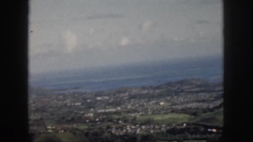1959 Oahu home-movie frame with a soft high view of coastal settlement, greenery, ocean, and hazy clouds.