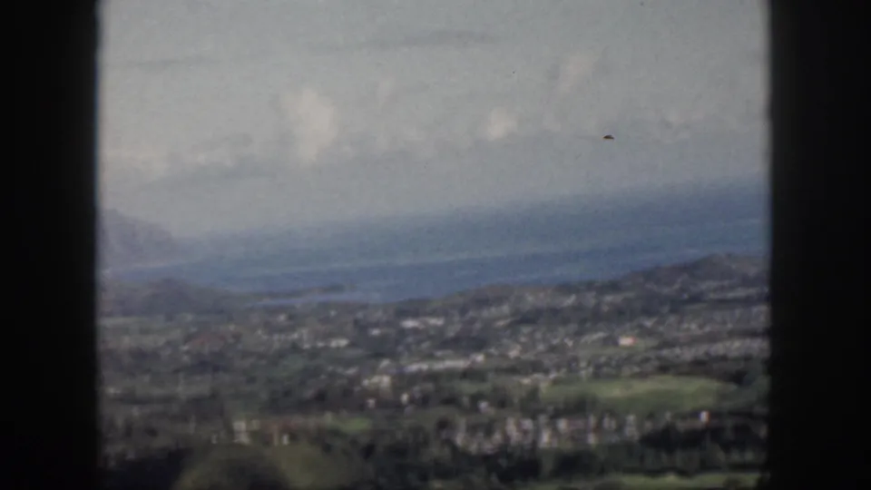 1959 Oahu overlook showing town, green hills, ocean water, distant landforms, and window framing in soft 8mm color.