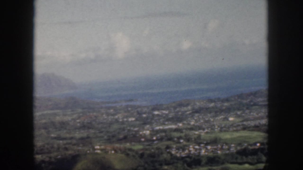 1959 Oahu home-movie frame showing a hazy airplane-window view over a coastal settlement, green hills, blue water, and distant mountains.
