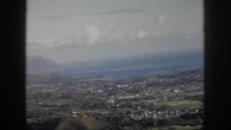 1959 Oahu home-movie frame showing a hazy airplane-window view over a coastal settlement, green hills, blue water, and distant mountains.